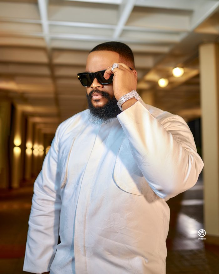 Whitemoney in a white agbada, hand to his sunglasses, in a lit corridor