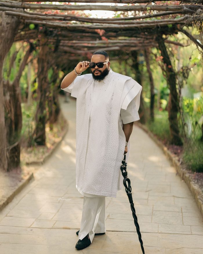 Whitemoney walking a garden pergola in a white agbada with a cane